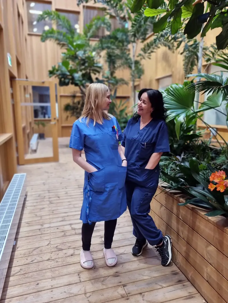 Två kvinnor i sjuksköterskekläder ler mot varandra i ett orangeri med gröna växter.